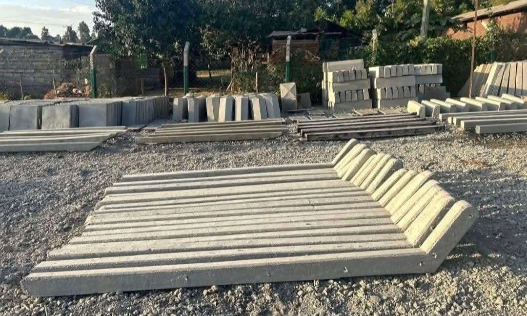 A fan of ridged concrete posts laid flat across the gravel yard, with stacks of road kerbs, slabs on edge and pillar caps lined up along the boundary wall behind them.