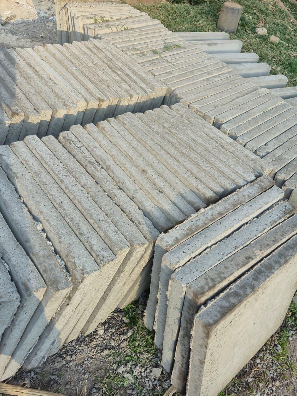 Flat slabs stacked in blocks on the gravel, most of them packed edge to edge so the stacks read as rows of close-set lines, with one slab at the front turned to show its plain face and a cut tree stump on the grass beyond.