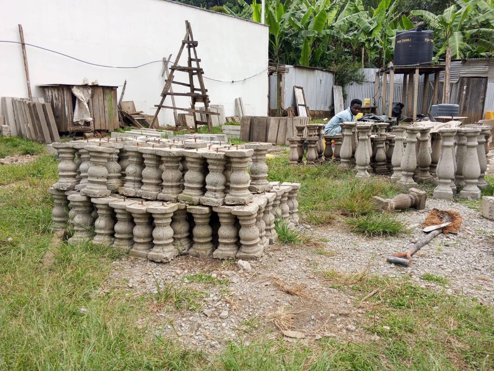 Rows of turned balusters stacked several courses high on the grass, each with a square cap and a steel fixing pin standing out of the top, while a worker sets out more of them on the gravel behind.