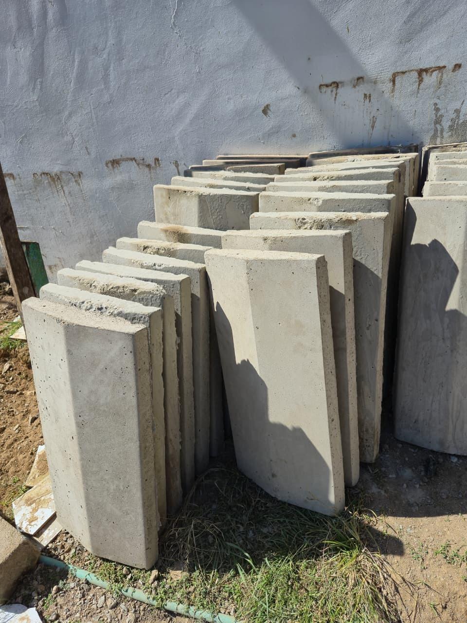 Slabs stacked on edge in the sun against a rendered wall, the nearest one turned to show its full face and the pitted texture of the cast surface.