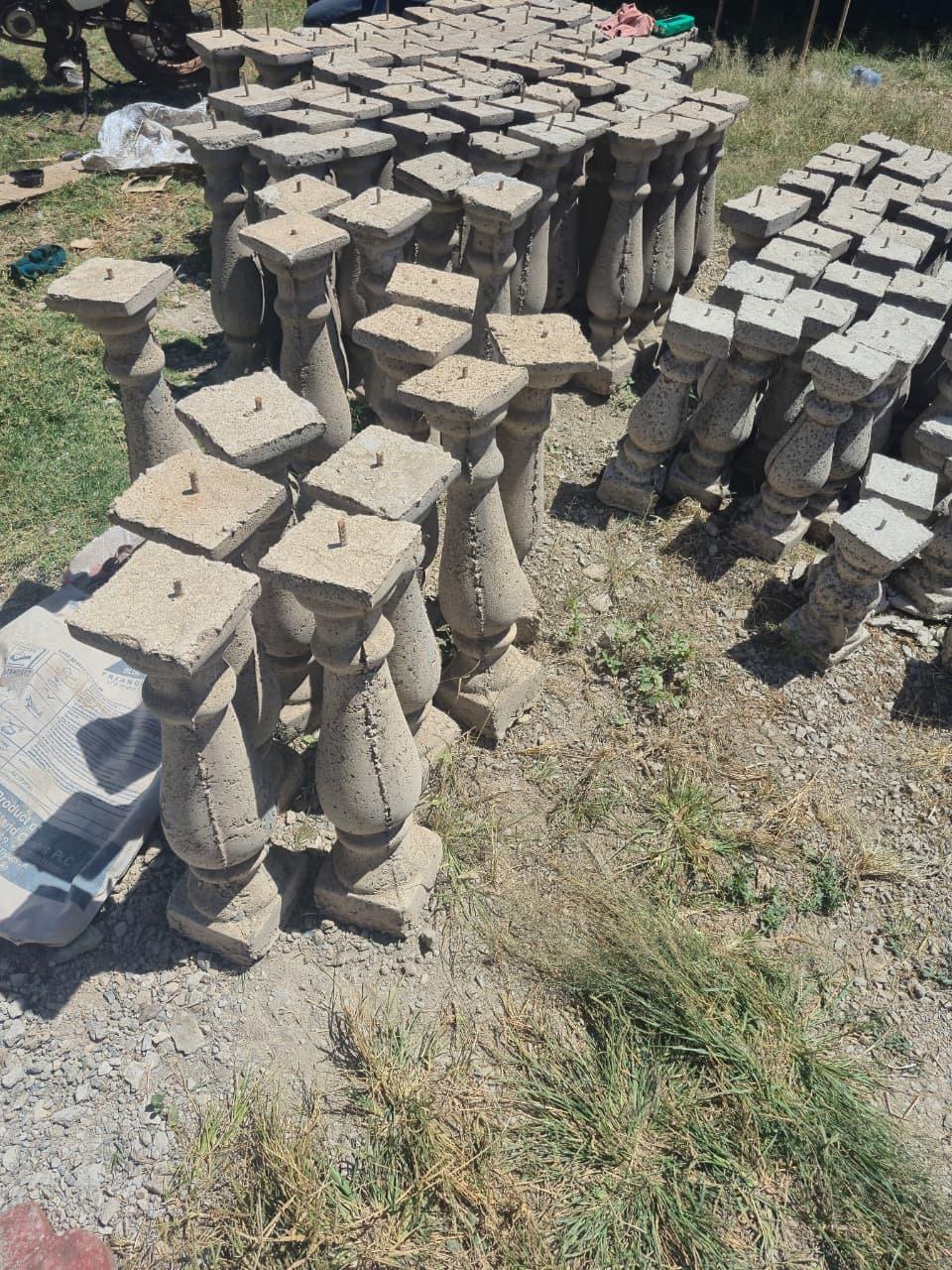 Balusters standing in tight blocks on the grass and gravel, seen from above so the square caps and their centre fixing pins fill most of the frame.