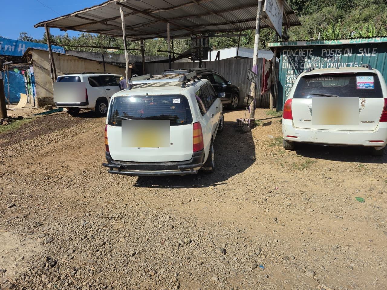 Cars parked on the gravel forecourt under the open iron-sheet canopy, with the green shop wall advertising concrete posts on the right.